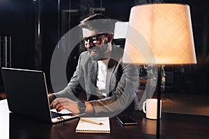 Smiling young bearded businessman working on contemporary notebook in loft office at night