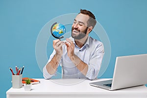 Smiling young bearded man in light shirt sit work at white desk with pc laptop isolated on pastel blue wall background