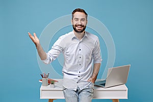 Smiling young bearded man in light shirt work near white desk with pc laptop isolated on pastel blue background