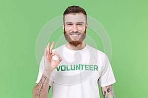 Smiling young bearded man in white volunteer t-shirt isolated on pastel green wall background studio portrait. Voluntary