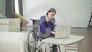 Portrait of a smiling young beautiful disabled woman in a wheelchair, working at home on a laptop, remote work