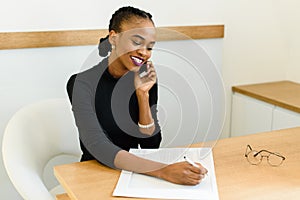 Smiling young black business woman on phone taking notes in office