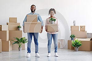 Smiling young black couple carry cardboard boxes, potted plants in room with white walls interior