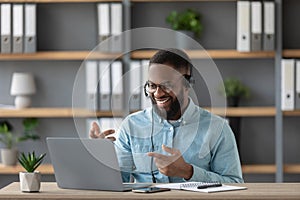 Smiling young black guy teacher or student with beard in glasses and headphones shows his fingers at pc screen