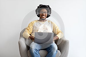 Smiling Young Black Guy Wearing Wireless Headphones Using Laptop Computer At Home