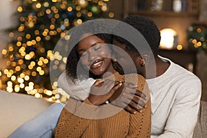 Smiling young black husband hug and kiss wife, enjoy holiday together in living room interior with Christmas tree