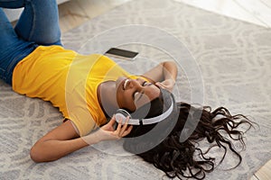 Smiling young black lady in headphones listening to music or audio book on floor at home