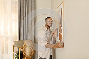 Smiling young black man putting picture frame, hanging painting on wall. Home interior and domestic decor