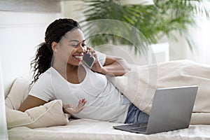 Smiling young black woman with cellphone and laptop relaxing in bed