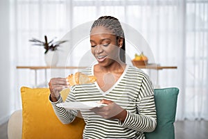 Smiling young black woman having breakfast at home, eating croissant