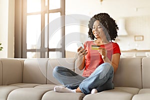 Smiling young black woman holding credit card and using smartphone at home