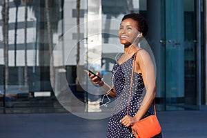 Smiling young black woman walking with earphones and mobile phone