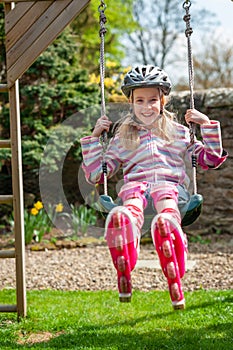 A smiling young blonde girl wearing roller blades and helmet while swinging on a garden swing