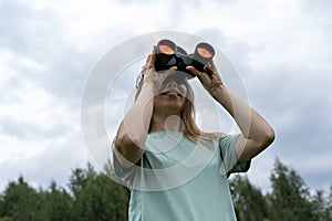 Smiling Young blonde woman bird watcher in cap and blue t-shirt looking through binoculars at cloudy sky in summer forest