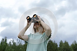 Smiling Young blonde woman bird watcher in cap and blue t-shirt looking through binoculars at cloudy sky in summer forest