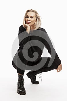 Smiling young blonde woman in black casual suit is squatting. White background. Vertical