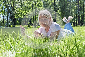 A smiling young blonde woman lies on the grass in a park with a laptop on a summer sunny day. Blogging, online communication,