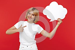 Smiling young blonde woman pointing finger on blank speech bubble over red background.