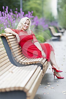 Smiling young blonde woman in a red dress sits on a bench in the park. Vertical