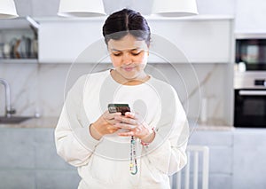 Smiling young brunette chatting on smartphone in home kitchen