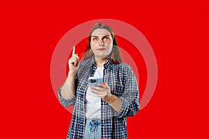 Smiling young brunette woman with smart phone pointing with the index finger a great idea on red background, studio