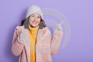 Smiling young brunette woman yellow sweater, pink fur coat , gray cap and mittens, posing isolated on lilac wall, pointing index
