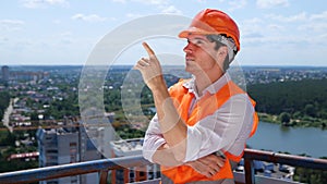 Smiling young builder in helmet pointing finger at construction site while standing on the roof. Business, building