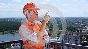 Smiling young builder in helmet pointing finger at construction site while standing on the roof. Business, building