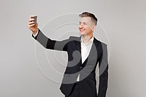 Smiling young business man in classic black suit, shirt doing taking selfie shot on mobile phone isolated on grey