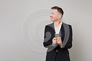 Smiling young business man in classic black suit, shirt looking aside using mobile phone typing sms message isolated on