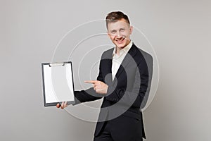 Smiling young business man in suit pointing index finger on clipboard with blank empty sheet workspace isolated on grey