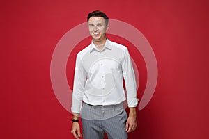 Smiling young business man in white shirt, gray pants posing isolated on bright red wall background studio portrait