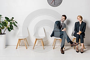 smiling young business people holding paper cups and looking away while waiting on chairs