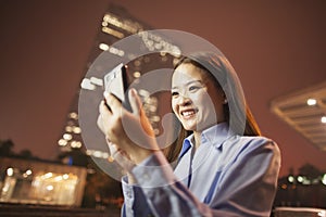 Smiling young business woman looking at her mobile phone outside at night