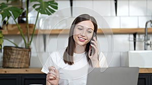 Smiling young business woman professional talking on phone using laptop at home, happy female customer make mobile call