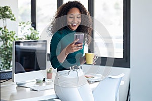 Smiling young business woman sending messages with mobile phone sitting on the desk in the office
