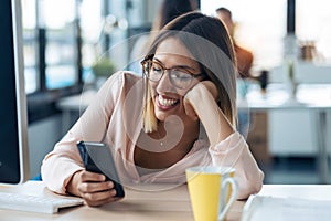 Smiling young business woman sending messages with mobile phone while sitting in modern startup office