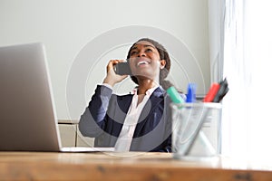 Smiling young business woman sitting at office desk and talking on cell phone