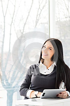 Smiling young business woman using tablet PC while standing relaxed near window at her office