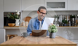 Smiling young businessman analyzing report and working over laptop on desk in home office