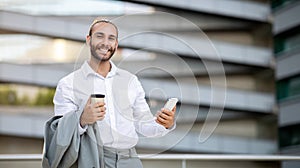 Smiling young businessman with coffee and phone posing outdoors