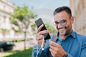 Smiling young businessman connecting headphones to smart phone at the city