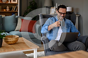 Smiling young businessman drinking coffee and reading e-mails over wireless computer while sitting on armchair. Happy male