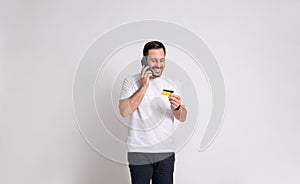 Smiling young businessman holding credit card and talking over smart phone against white background