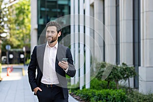 Smiling young businessman man in a business suit is using a mobile phone on the street, walking on a break outside