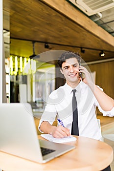 Smiling young businessman sitting behind his desk with laptop and talking on mobile phone in office