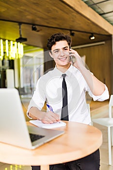 Smiling young businessman sitting behind his desk with laptop and talking on mobile phone in office