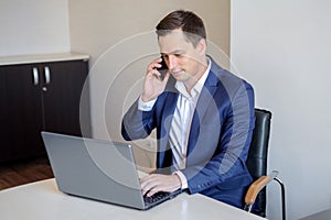 Smiling young businessman sitting behind his desk with laptop and talking on mobile phone in office