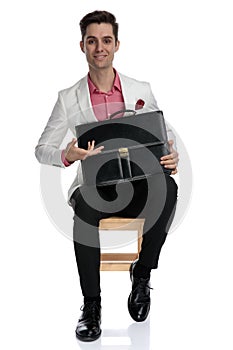 Smiling young businessman sitting and presenting his briefcase