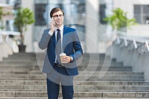 smiling young businessman in stylish suit with coffee to go talking by phone on stairs